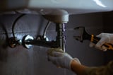 Someone in white gloves works on plumbing under a white sink using an adjustable wrench